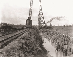 A crane driving piles into the ground moves across railroad tracks. 