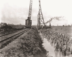 A crane driving piles into the ground moves across railroad tracks. A crane driving piles into the ground moves across railroad tracks.