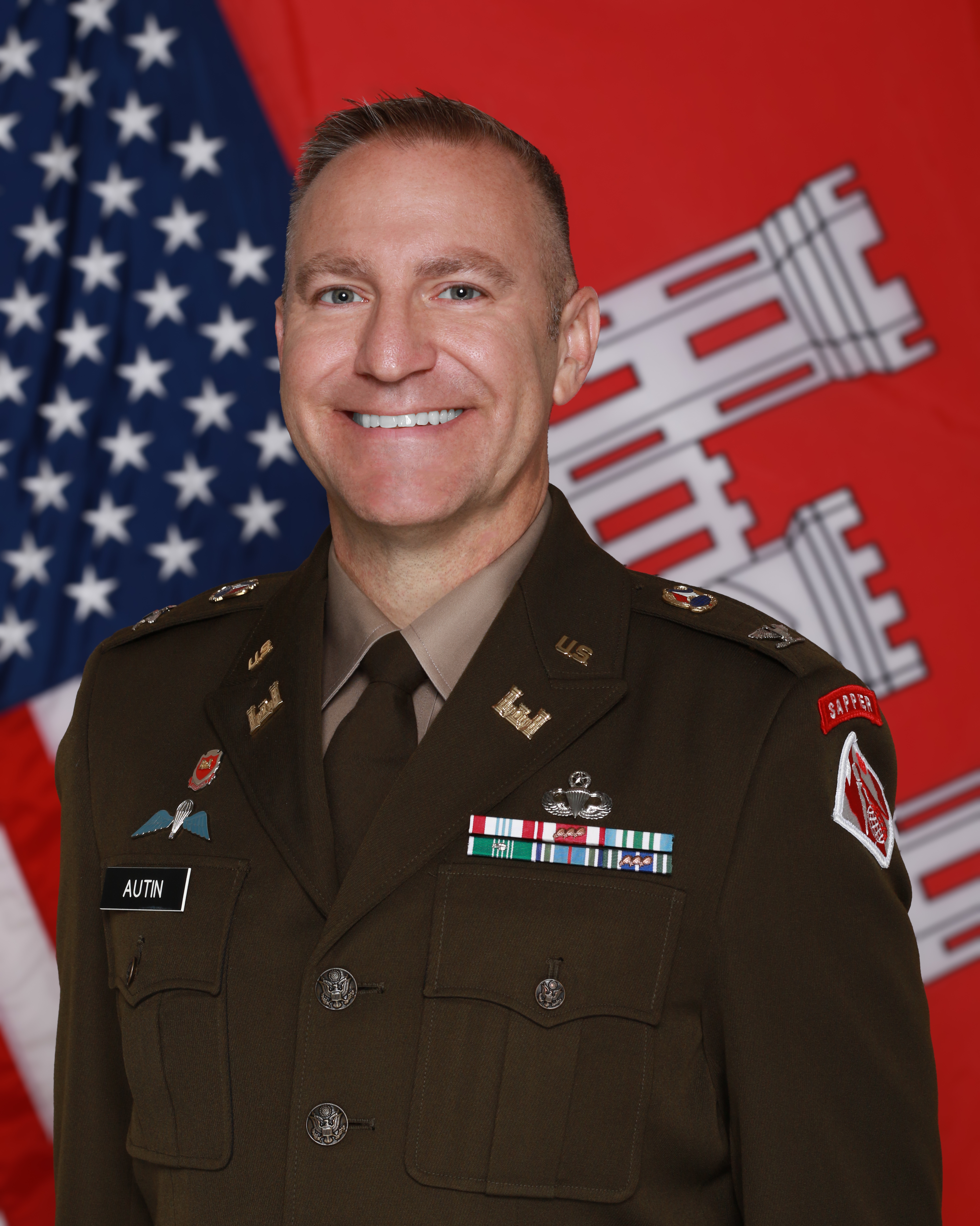 Colonel Autin, wearing the Army Green Service Uniform, stands in front of the American and U.S. Army Corps of Engineers flags.